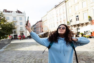 Cheerful curly haired woman traveler wearing sunglasses taking s clipart