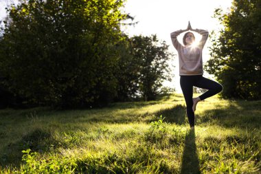 Yoga yapan kadın parkta Vrikshasana pozu veriyor. Gün batımında kadın silueti. Canlılık kavramı, denge, farkındalık, sakinlik, rahatlama..