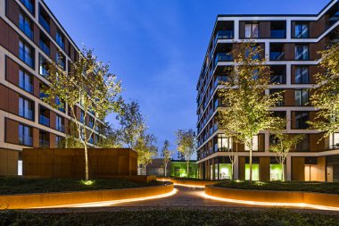 Geceleri bir Avrupa şehrinde konut binaları. Modern apartman blokları. Bahçede bitki örtüsü ve ışıklandırma var. Paslı metal kaplama, korten. Yeraltı garajı