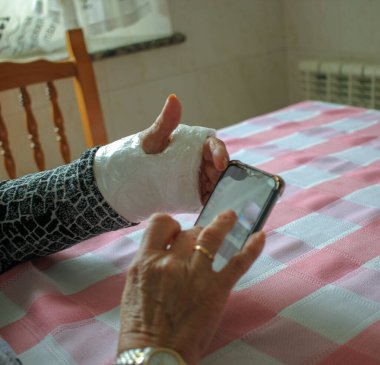 Ailesiyle iletişim kurmak için cep telefonuna bakan kollu yaşlı bir kadın.