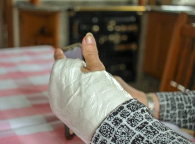 Bileği alçılı yaşlı bir kadın evde cep telefonunu izliyor.