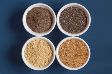 Top view of 4 food bowls filled with chia and flax both ground and whole on a blue background. Seeds and food.