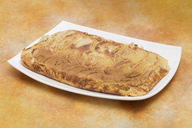 Üzerinde tarçın olan güzel pişmiş elmalı turta, beyaz, dikdörtgen bir tabakta altın kahverengi bir kabuk ve içinde elma dilimleri ve sıcak turuncu bir arka plan ile sunuluyor.. 