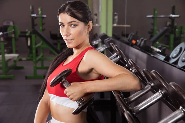 Kırmızı beyaz eşofmanlı güzel esmer, spor salonunda dambılları kaldırıyor. Kırmızı askılı uzun pembe tırnaklı seksi esmer ve spor salonundaki halterler ve ağırlıklar arasında taytlı.
