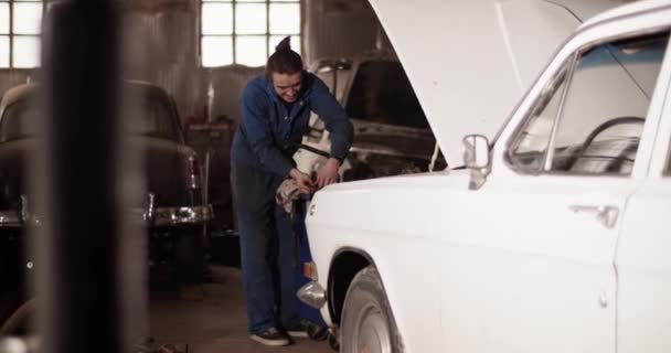 ガレージで白いレトロな車を修理しながら インストール前にシリンダーヘッドカバーを検討作業服の全長技術者の手持ち撮影 — ストック動画