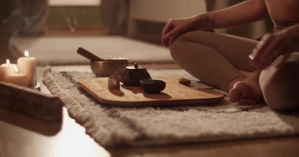 在家庭瑜伽课上 匿名的女性双腿交叉坐在垫子上 将传统的中国茶倒入芬芳的蜡烛和熏香旁的杯子中 — 图库视频影像