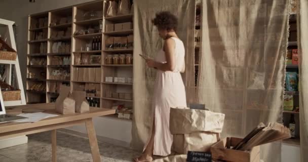 用平板电脑拍摄穿着白色衣服的黑人女性手持照片 检查网上客户的订单 同时从有机产品商店的货架上挑选蜂蜜 — 图库视频影像