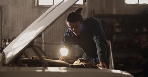 ガレージでの車両検査中に壊れた車のエンジンを照らすために懐中電灯を使用して若い男のハンドヘルドショット — ストック動画