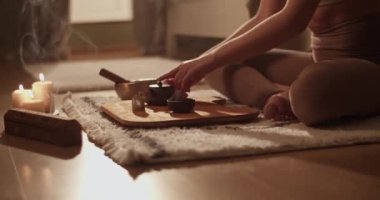 Anonim bir kadın paspasın üzerinde oturur ve evde yoga sırasında aromatik mumlar ve tütsü eşliğinde geleneksel Çin çayını bardağa doldurur.