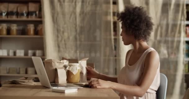 アフリカの髪を持つ若い民族の女性は オーガニック製品とテーブルに座って エコグッズの自分の店でノートパソコンで作業しながら出荷のためのアイテムを準備 — ストック動画