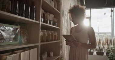 Afro saçlı genç bir kadın dükkandan sipariş vermek için tablet kullanırken raftan bal alıyor güneşli bir depoda çalışıyor.
