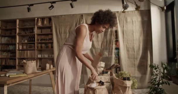 エコ製品で店で働くオンライン購入者の注文を梱包しながら 紙袋に穀物を注ぐドレスの若い民族の女性 — ストック動画