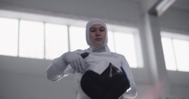 Gün ışığında güneşli spor salonunda eğitime hazırlanırken folyo giymiş, koruyucu maske takmış ve kollarını kavuşturmuş Müslüman kadın eskrimcilerin düşük açılı el kamerası görüntüleri.