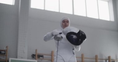 Gün ışığında güneşli spor salonunda eğitime hazırlanırken folyo giymiş, koruyucu maske takmış ve kollarını kavuşturmuş Müslüman kadın eskrimcilerin düşük açılı el kamerası görüntüleri.
