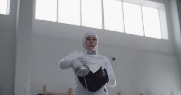 昼間の日光のジムでのトレーニングの準備中に保護マスクや折り畳み式の腕に置く箔と一様でイスラム教徒の女性フェンサーの低角度ハンドヘルドショット — ストック動画