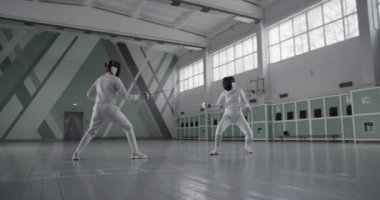 Üniformalı ve maskeli, tanınmayan kadın eskrimcilerin, geniş spor salonundaki antrenmanlar sırasında yaptıkları düelloyu izliyoruz.
