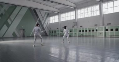Üniformalı ve maskeli, tanınmayan kadın eskrimcilerin, geniş spor salonundaki antrenmanlar sırasında yaptıkları düelloyu izliyoruz.
