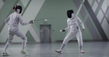 Koruyucu üniformalı, folyolu bir kadın sporcunun spor salonundaki eskrim antrenmanı sırasında rakibiyle pratik yaparken görüntüsünü yakınlaştır.