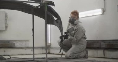 Kapaklı iş kıyafetleri ve solunum cihazıyla çalışan kadın boyacının el kamerasıyla çekilmiş bir fotoğrafı. Garaj atölyesinde çalışırken dizlerinin üzerine çöküp tampona boya sıkarken.