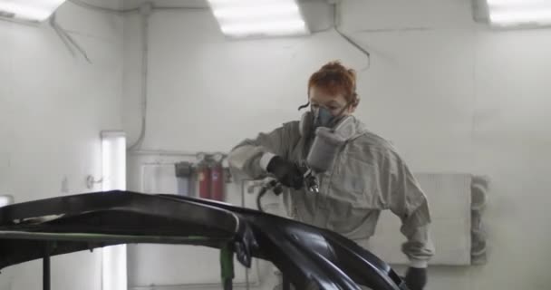 ガレージでの作業中にスプレー塗料で車のバンパーをカバーする全体的な均一でマスクで赤毛の女性の整備士のハンドヘルドショット — ストック動画
