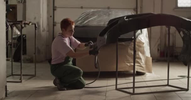 車の近くの床にひざまずいて全身の女性技術者のハンドヘルドショットと昼間の日光のワークショップでの作業中に絵のためのバンパーを準備 — ストック動画