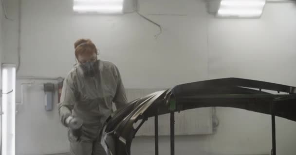 ガレージでの照明ワークショップでの作業中に黒い車のバンパーに塗料を噴霧全体の均一な呼吸器で職人の女性のハンドヘルドショット — ストック動画