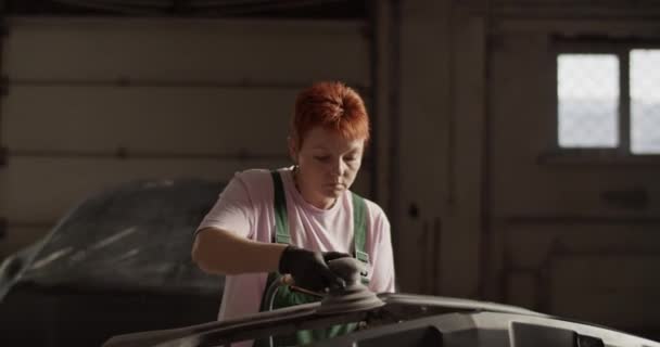 プロのガレージで作業しながら 車の近くのラック上の均一なサンディングバンパーに生姜女性の整備士のハンドヘルドショット — ストック動画