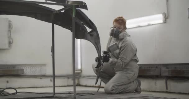 ガレージのワークショップ中に床にひざまずき バンパーに塗料を噴霧し 全体の作業服やマスクで全身の女性画家の手持ち撮影 — ストック動画