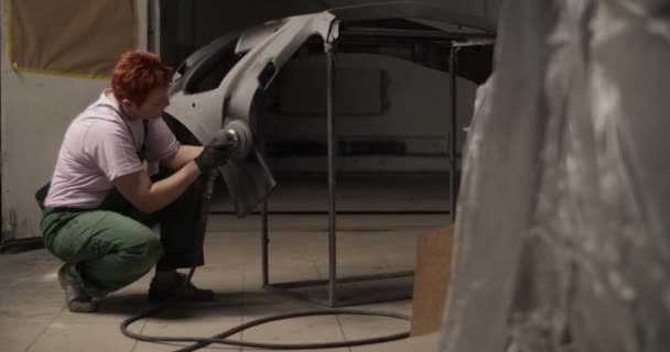 日光ガレージでの作業中にベンチに座って車のバンパー上のサンディングマシンを使用して修理女性の手持ち撮影 — ストック動画