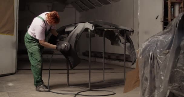 昼間のガレージ作業中にフィルムで覆われた車の近くのバンパー上のサンディングマシンを使用して均一で全身の女性技術者の手持ち撮影 — ストック動画