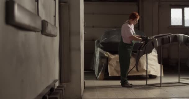 ガレージでの作業中に車の近くに立つ作業着とラックの上にバンパーをサンディングで全身の女性技術者の手持ち撮影 — ストック動画