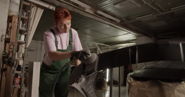 絵のワークショップで作業しながら バンパー上のサンディングマシンを使用して赤い髪で作業服の女性の低角度ハンドヘルドショット — ストック動画