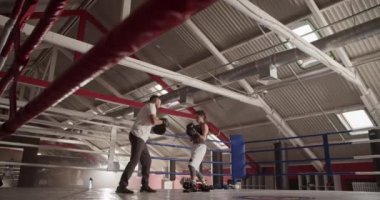 Düşük açılı el kamerasıyla, kadın boksörün, erkek antrenörle güneş ışığı altındaki spor salonunda antreman yaparken yumruk ve kaçamak hareketleri çekmesi.