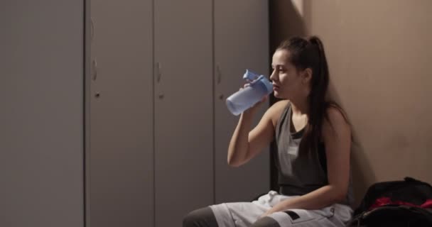 ジムのドレッシングルームでのワークアウトの後 ベンチに座って水を飲む疲れ女性選手のハンドヘルドショット — ストック動画