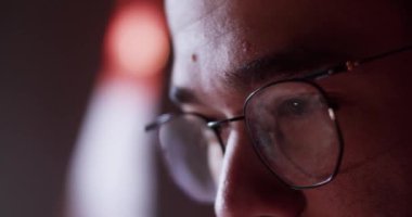 Closeup of cropped guy in glasses focusing on screen while playing videogame in dark room