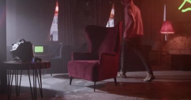 Casual male in hoodie entering room and taking gamepad from table with small TV and sitting in wingchair while lounging in hookah bar