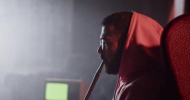 Side view of millennial male in glasses and hoodie sitting in armchair and blowing smoke rings of hookah while having cool drink in dark lounge bar