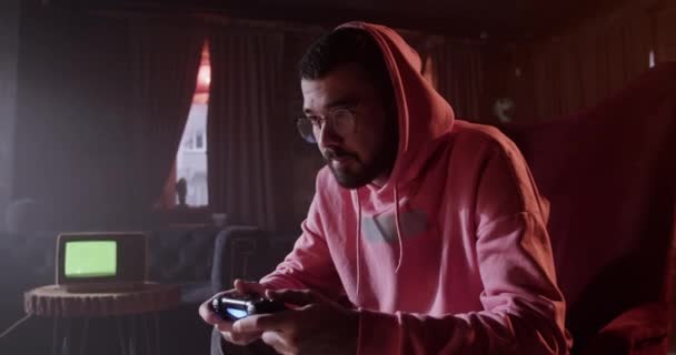 ミレニアム男性でパーカーと眼鏡でアームチェアに座っている薄暗いカフェやビデオゲームだけで遊ぶことに焦点を当てて — ストック動画