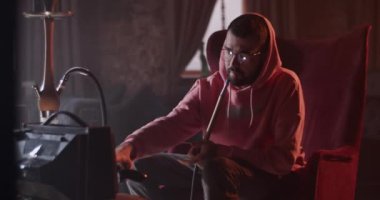 Young guy in hoodie putting gamepad on table while taking break and drinking cool beverage having hookah to chill in dark bar