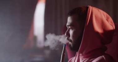 Side view of bearded millennial man in hoodie exhaling smoke of hookah and blowing smoke rings in dark bar