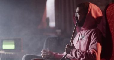 Casual young man sitting in armchair in lounge bar with glass of whiskey and blowing smoke rings while smoking hookah in dark