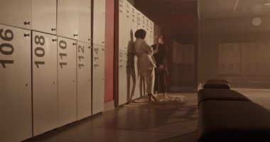 Handheld shot of barefoot diverse women tying belts on gis and talking with each other while standing near lockers and preparing for grappling training in gym