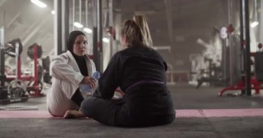 Handheld shot of happy multiethnic women drinking water and talking while sitting on floor during break in Brazilian jiu jitsu training in gym