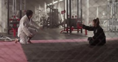 Handheld shot of Muslim wrestler doing headstand drill near female instructor sitting cross legged on floor during Brazilian jiu jitsu training behind net in gym