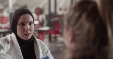 Handheld shot of young woman in white gi and black hijab talking with female coach during grappling training in gym