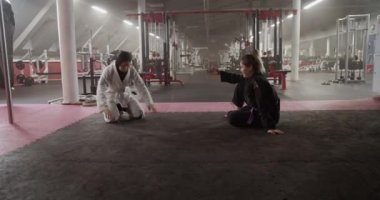 Handheld shot of Muslim wrestler doing headstand drill near female instructor sitting cross legged on floor during Brazilian jiu jitsu training behind net in gym