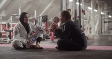 Handheld shot of happy multiethnic women drinking water and talking while sitting on floor during break in Brazilian jiu jitsu training in gym