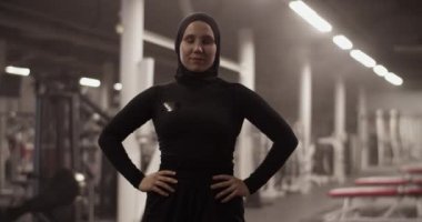 Handheld shot of strong Muslim female athlete in black sportswear and hijab holding hands on waist and looking at camera during workout in spacious gym