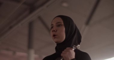 Low angle handheld shot of female athlete in black sportswear and hijab breathing and doing biceps curls with dumbbells during fitness workout in gym
