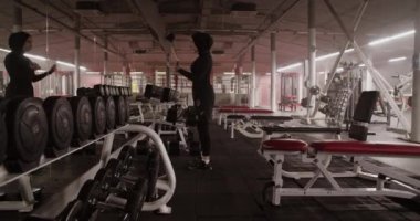 Handheld shot of Muslim sportswoman looking in mirror and doing biceps curls with dumbbells during weightlifting workout in spacious gym